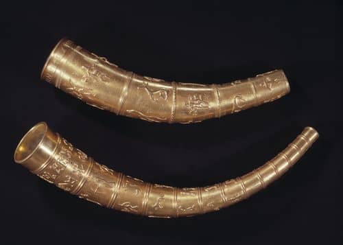 De to buede guldhorn med indgraverede figurer og mønstre ligger på en mørk baggrund. Det ene horn er tykkere med en bred åbning, mens det andet er smallere og længere.