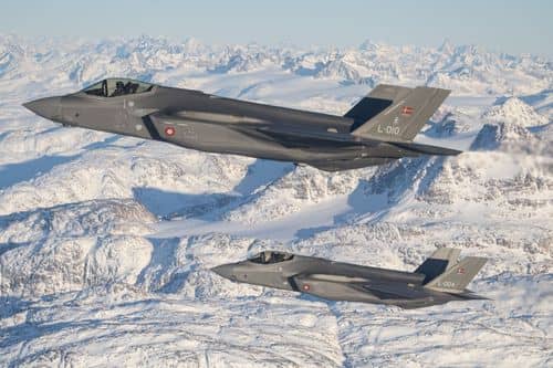 To danske F-35-kampfly flyver i formation over et snedækket bjerglandskab langs Grønlands østkyst. Billedet illustrerer Danmarks styrkede overvågning og militære tilstedeværelse i Arktis og Nordatlanten.